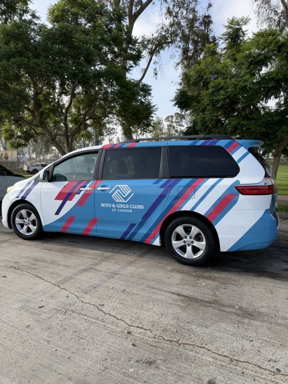 Wrapped white minivan with blue and red graphics parked outdoors under trees