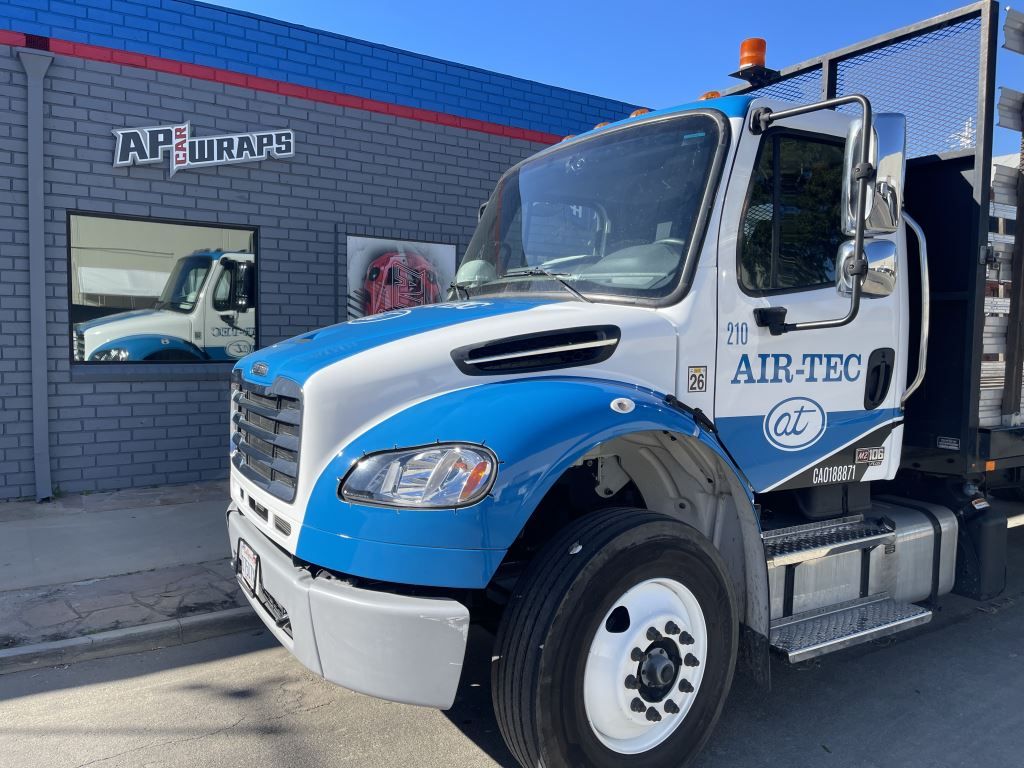 Blue and white utility truck parked outside a commercial building with a logo on the wall