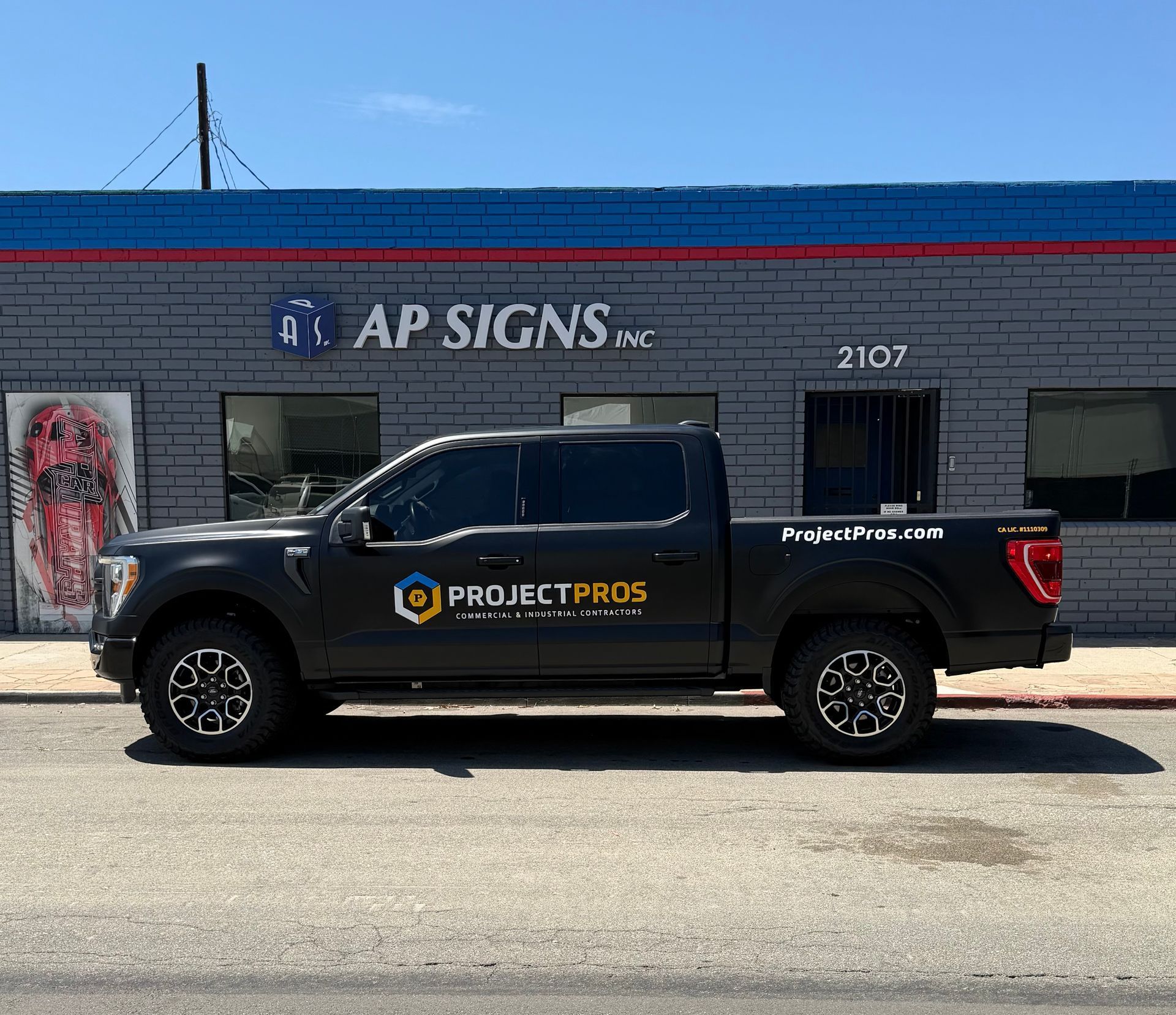 Black Project Signs pickup truck parked in front of an AP SIGNS storefront.