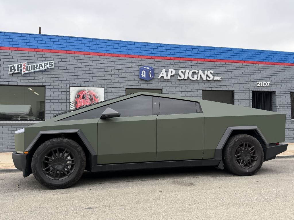 Matte green futuristic armored pickup parked outside an APS Signs storefront