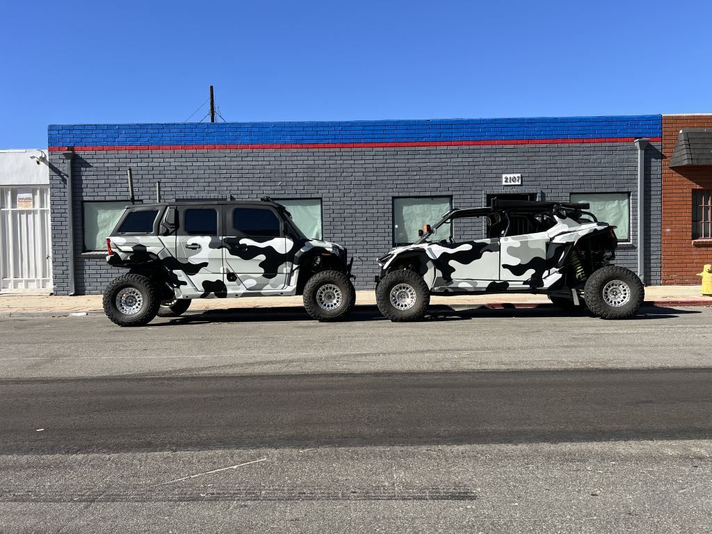 Two camouflaged off-road vehicles were parked side by side in front of a gray building.