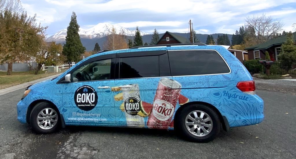 Blue minivan with large soda can graphics parked outdoors on a cloudy day