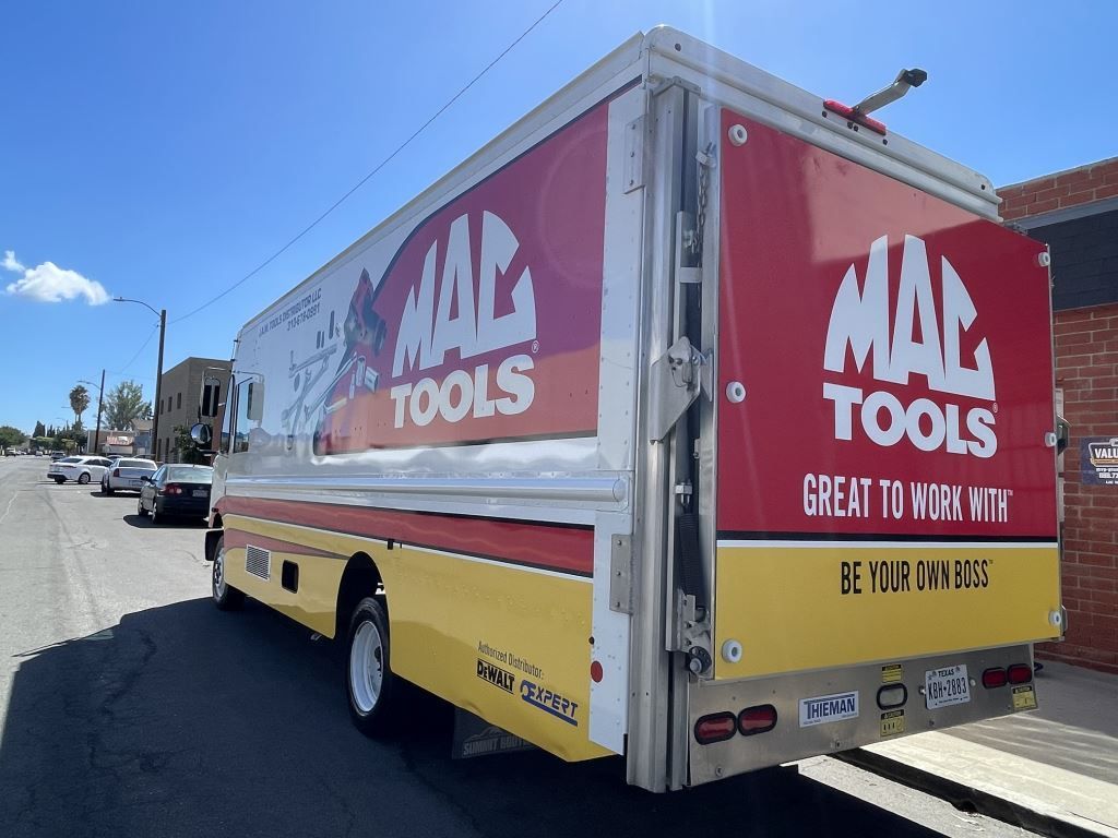 Mack Tools box truck parked on a street, red and yellow ad panels visible on the side and rear.