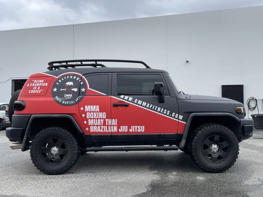 Black off-road SUV with red graphic wrap and roof rack parked by a plain white wall.