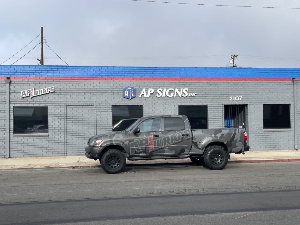 Camouflaged pickup truck parked outside AP Signs storefront with blue and red trim.