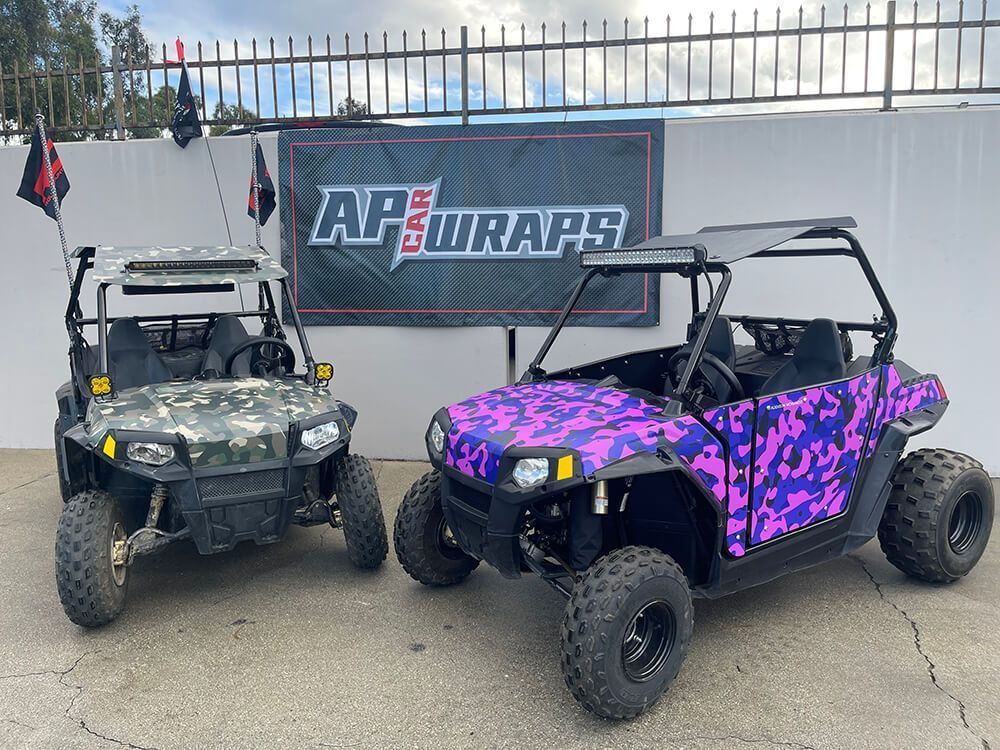 Two camo UTVs, one purple and one gray, are parked in front of an AF Wraps sign.