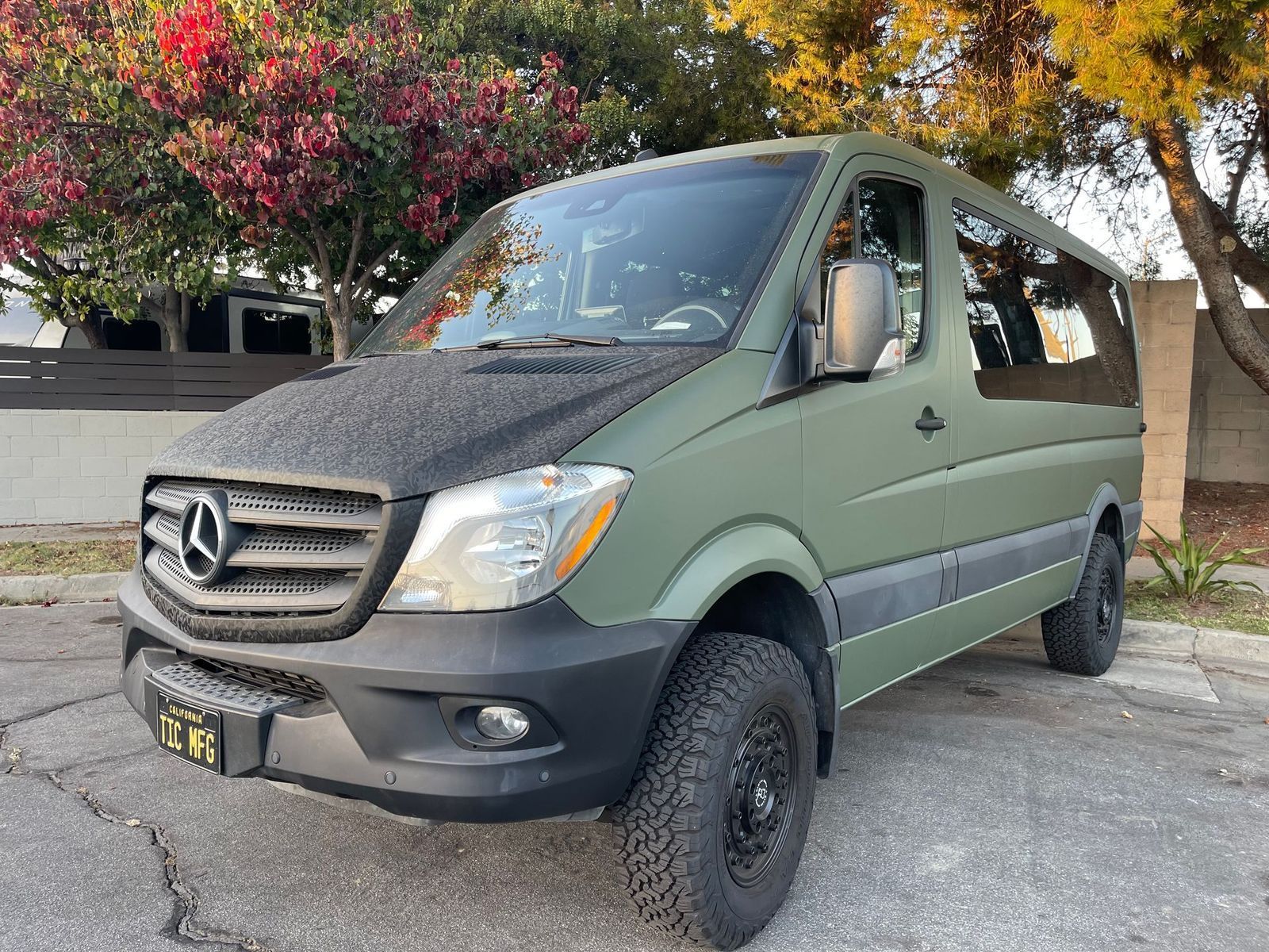 Green Mercedes van with off-road tires parked outdoors near trees and a building.