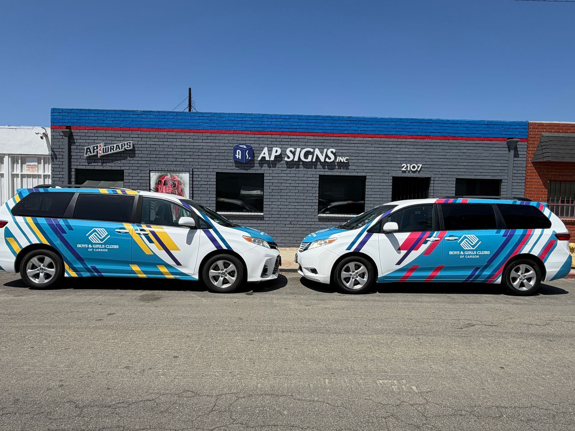 Two branded cars parked outside an AP Signs storefront, each wrapped in blue and pink graphics.