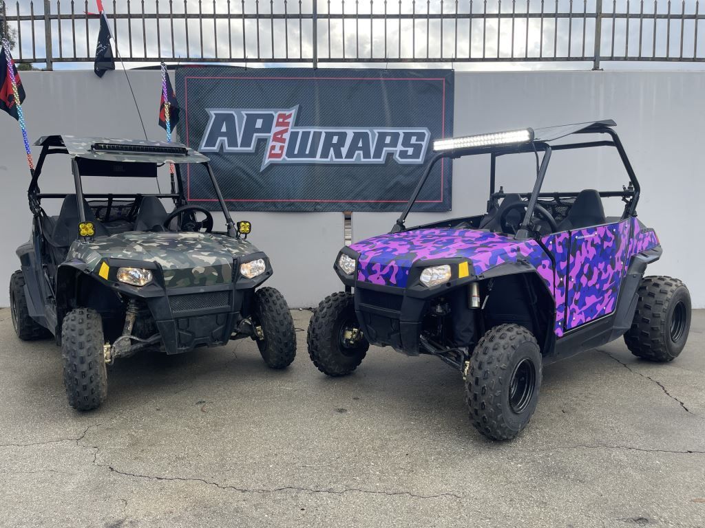 Two off-road dune buggies, one black and one purple camouflage, parked in front of an AP Wraps banner.