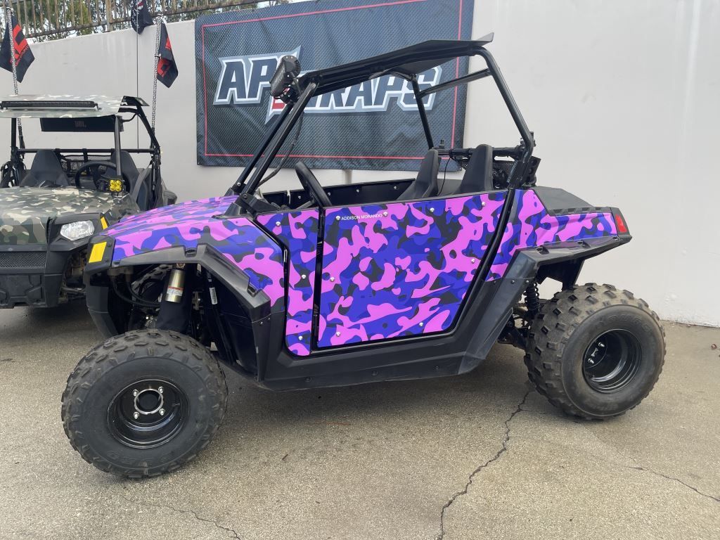 Purple camo off-road side-by-side UTV parked on pavement, with black roll cage and rugged tires.