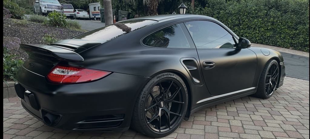 Matte black sports coupe parked on a cobblestone driveway beside greenery.