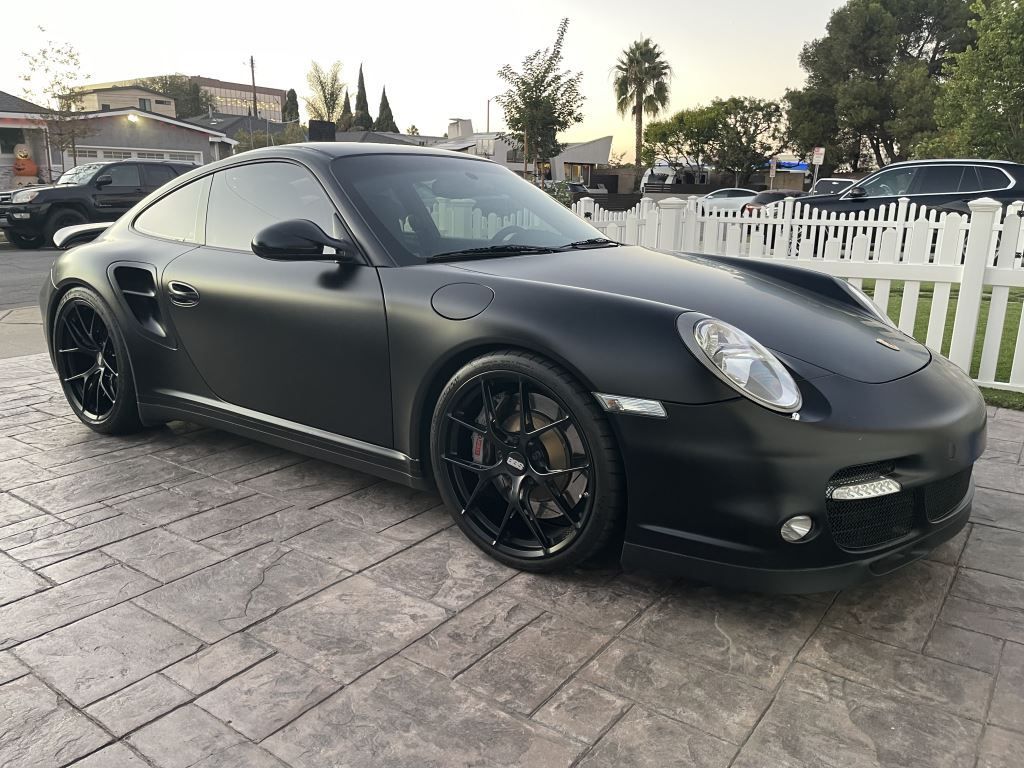 Matte black Porsche 911 parked on a driveway beside a white fence at dusk