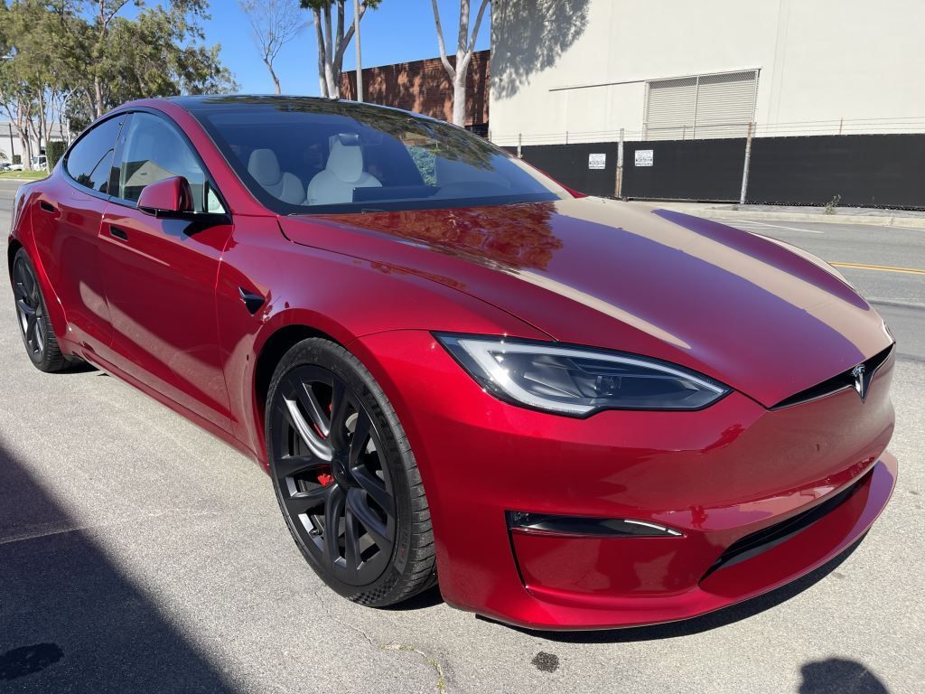 Red Tesla sedan parked outdoors in sunlight, with a glossy red finish and black wheels.