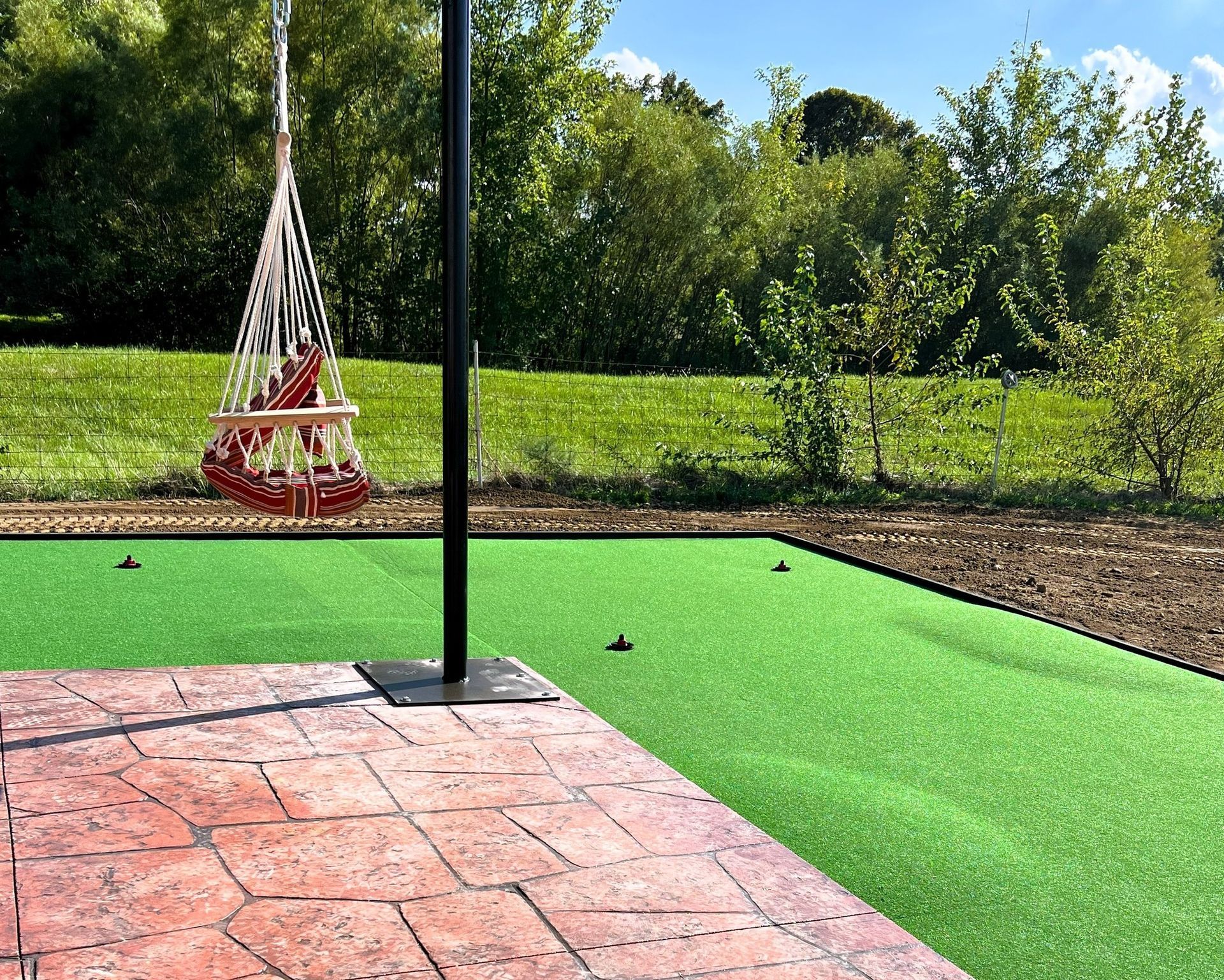 A backyard scene with a putting green, swing chair, and brick patio on a sunny day.