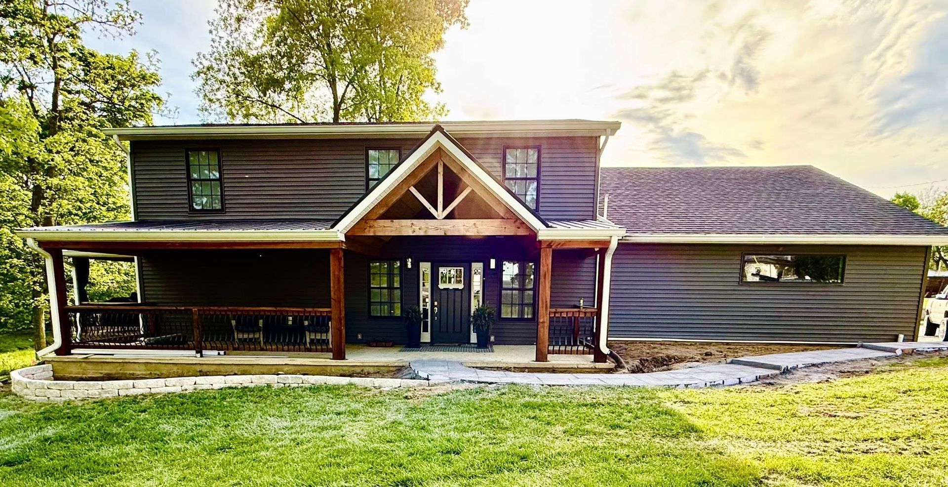 Two-story house with dark siding, brown porch and wooden accents, in a grassy setting with trees and blue sky.