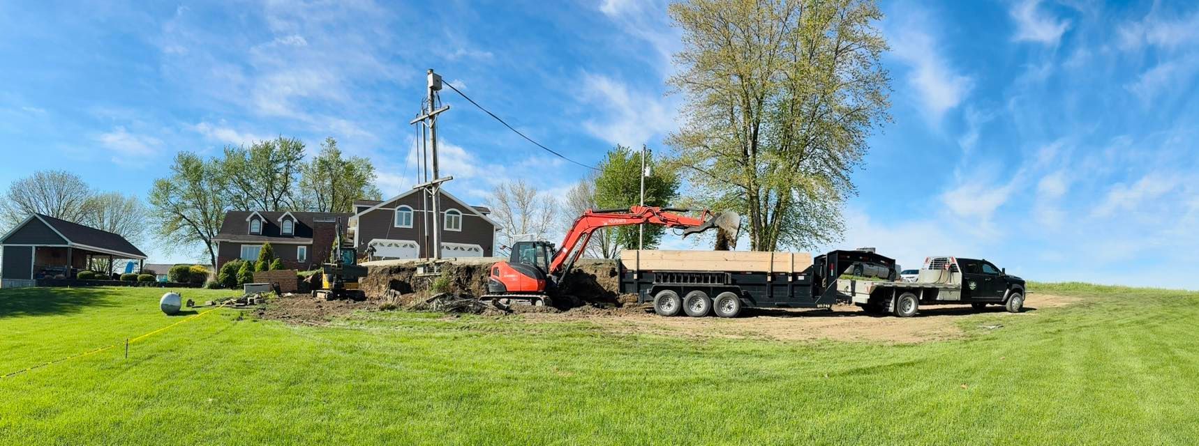 An excavator loading dirt onto a flatbed trailer beside a house. A truck is parked next to the trailer. Over a field of green grass and blue sky.
