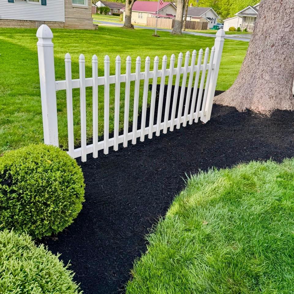 White picket fence surrounds a bed of black mulch next to a tree on a green lawn.