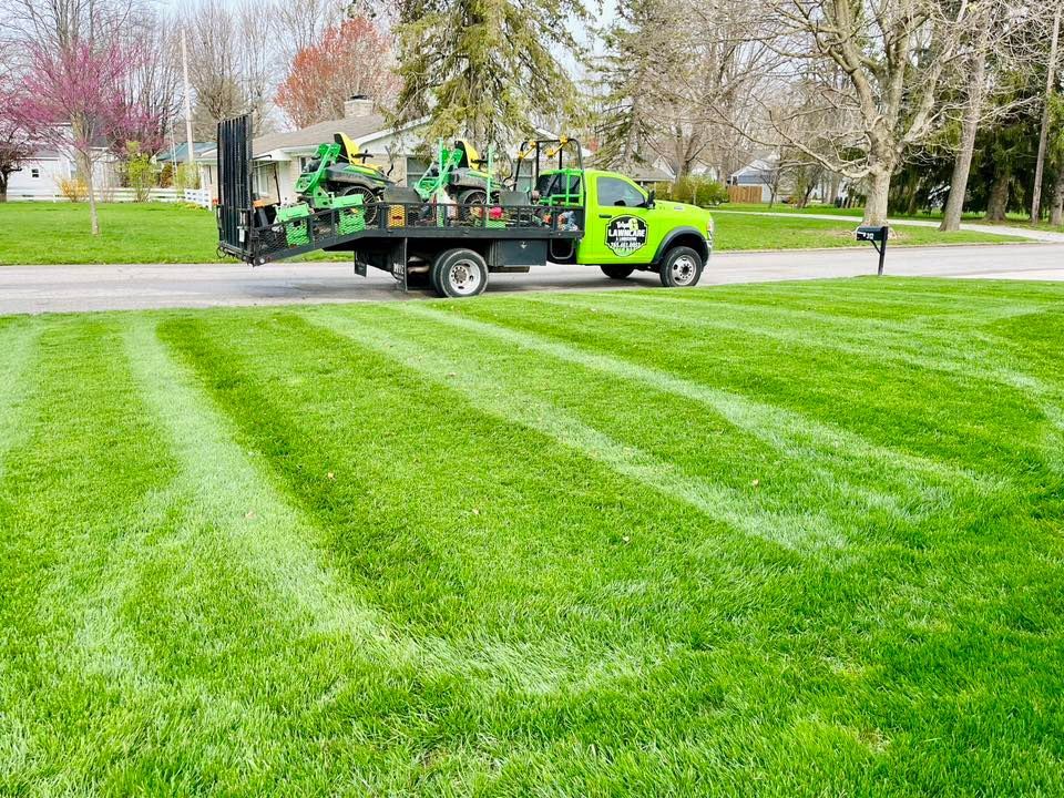 A green lawn care truck parked on a lawn with freshly mowed stripes, in a suburban neighborhood.