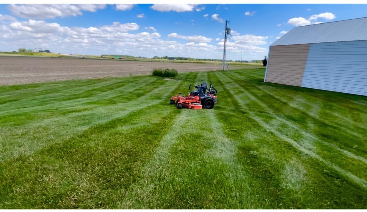 A tractor mowing a large lawn, creating stripes on the green grass under a partly cloudy sky.  A white building sits on the right.