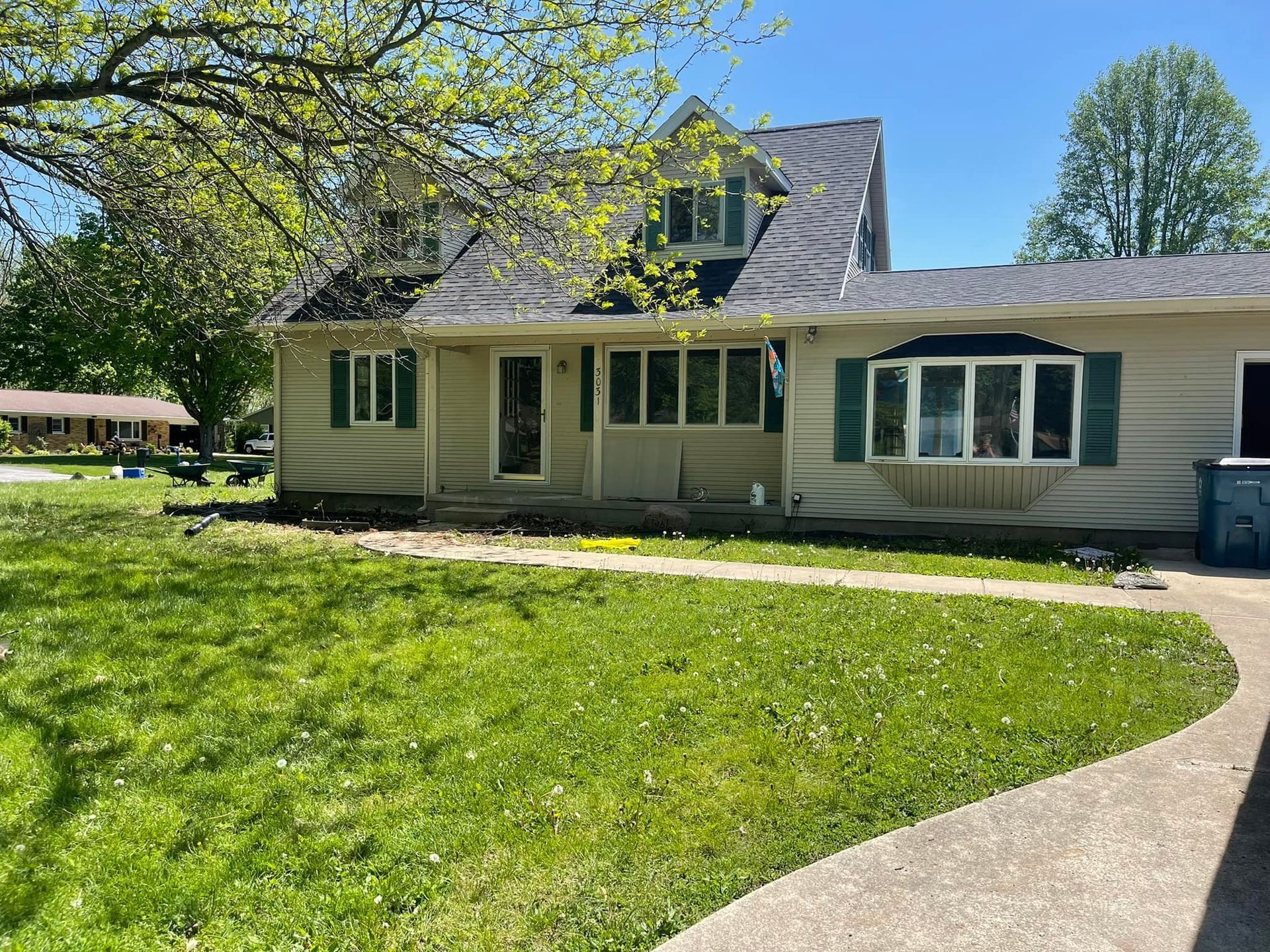 A light tan house with green shutters and a bay window. A sidewalk curves through the green lawn in front.