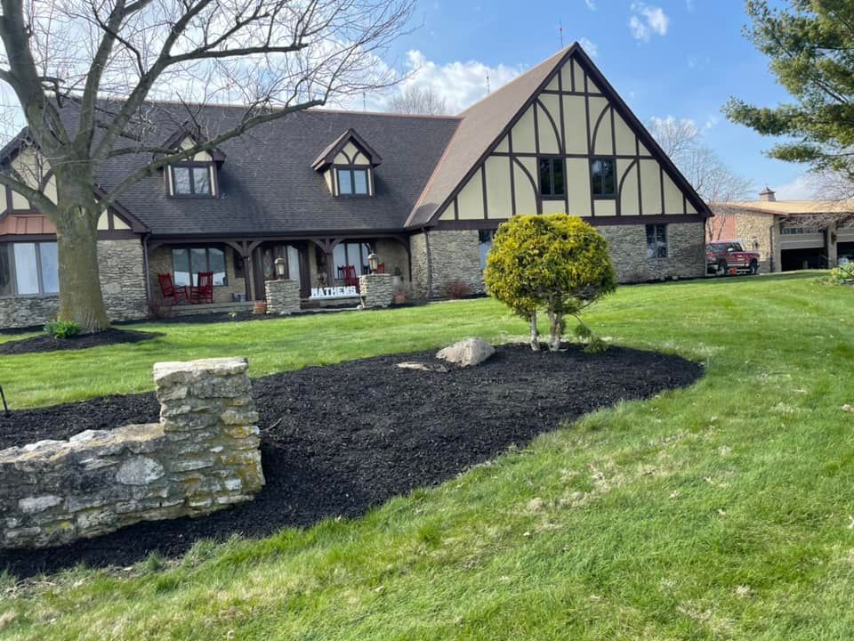 A two-story Tudor-style house with a stone facade and brown roof sits on a grassy lawn. Black mulch borders the front.