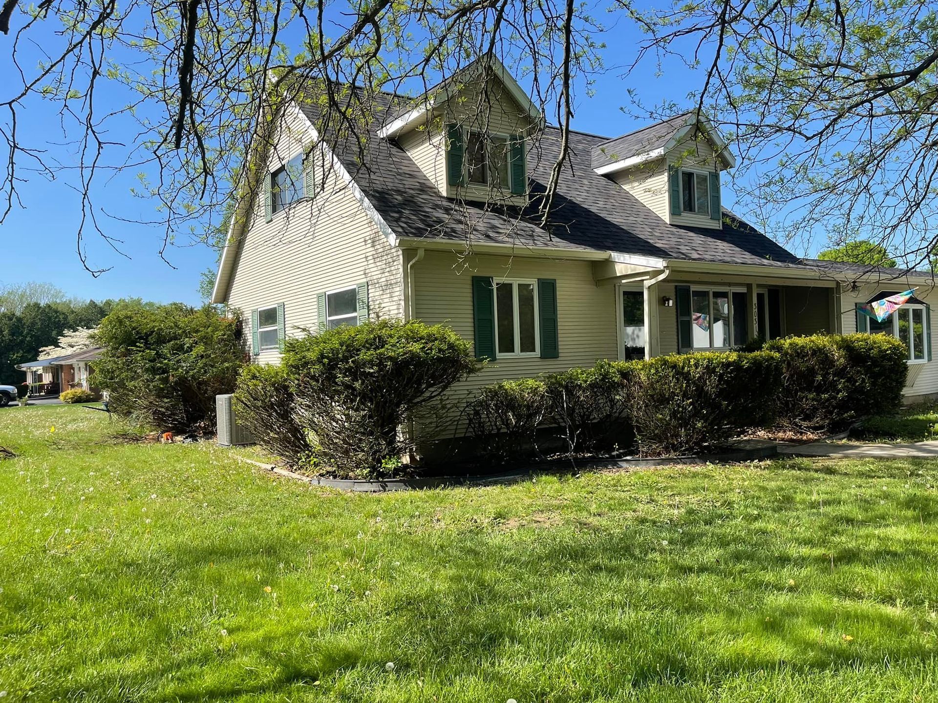 A two-story, beige house with green shutters and dormers, framed by tree branches, sits on a grassy lawn under a blue sky.
