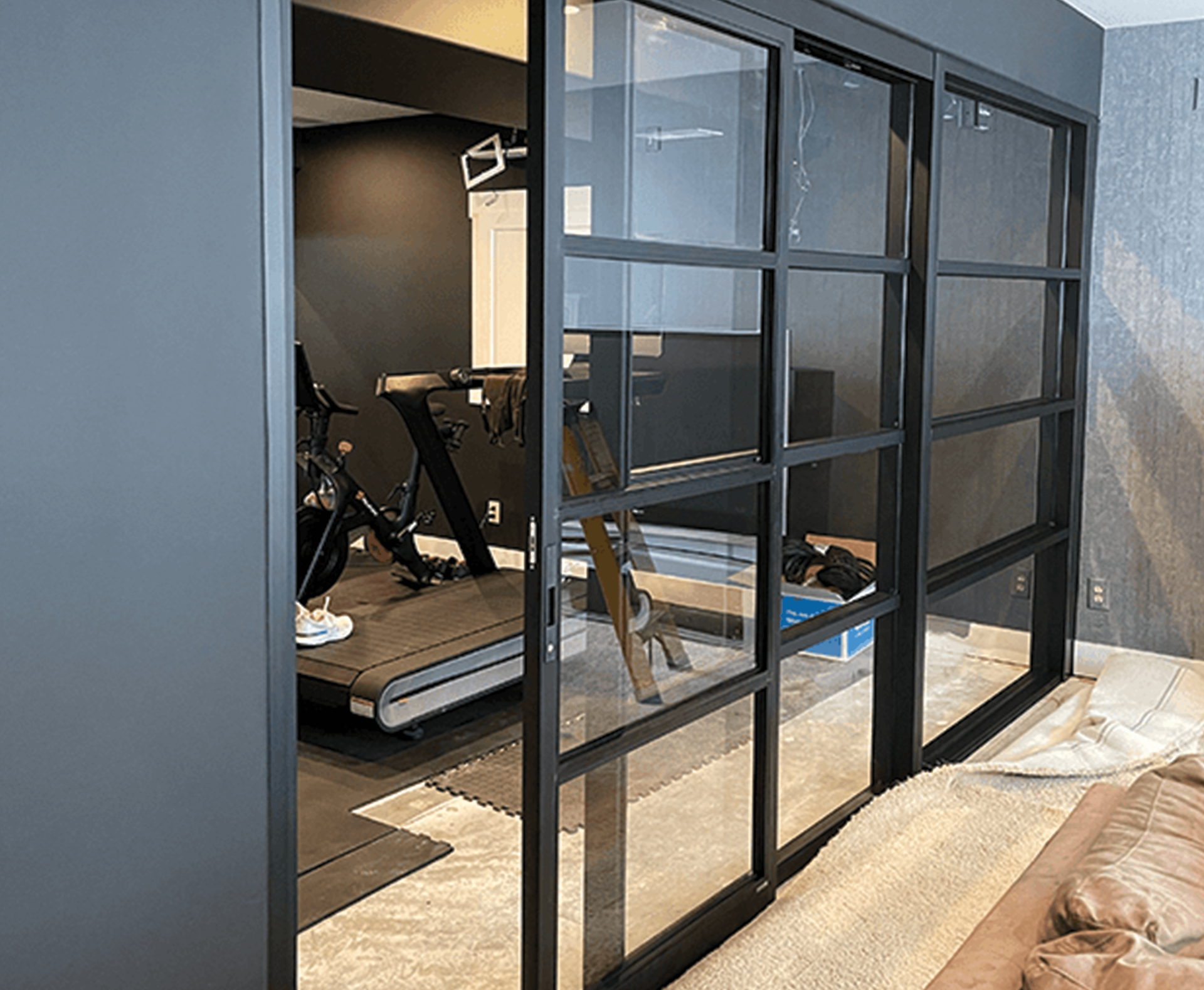 Black-framed glass sliding doors separate a bedroom from a home gym area with a treadmill and exercise bike.
