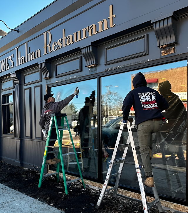 Two people cleaning windows of an Italian restaurant. Exterior view, gray building, sunny day.
