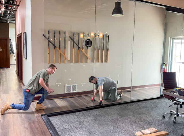 Two people installing a large mirror on a wall. One kneels, the other crouches, both focused. Interior setting.