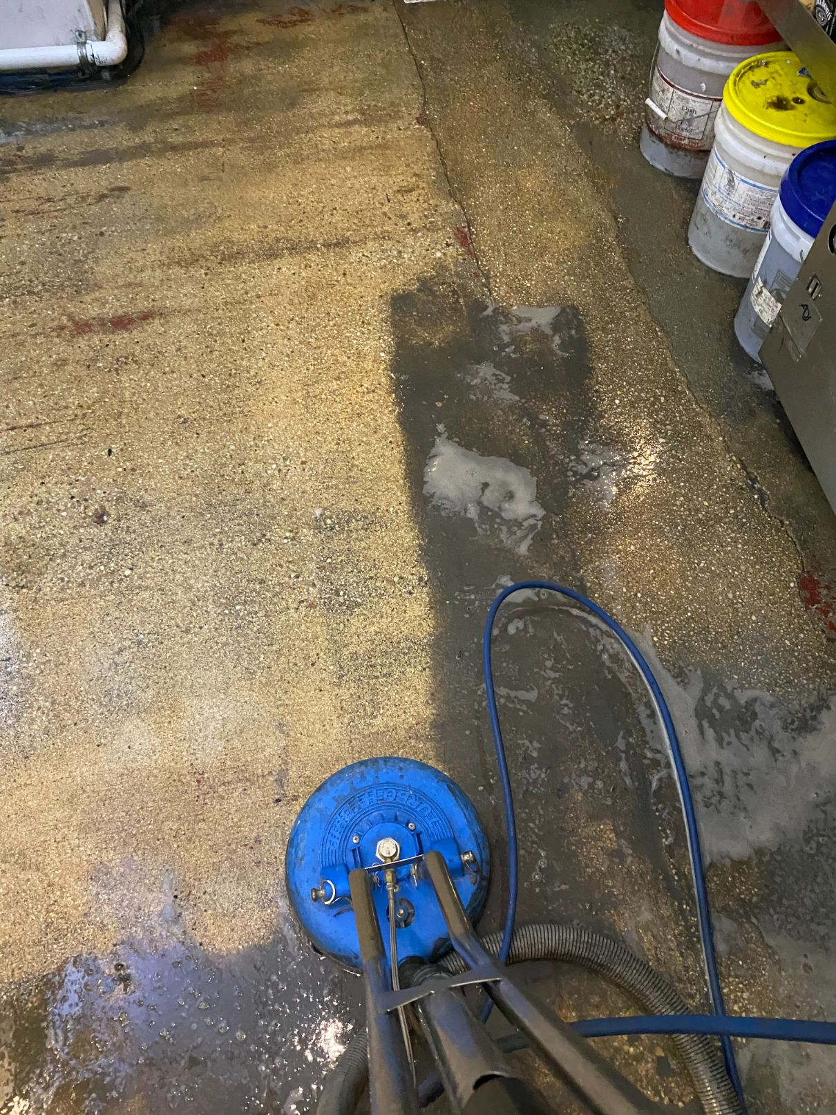 A blue surface cleaner scrubbing a wet, speckled concrete floor. Buckets are visible on the right.