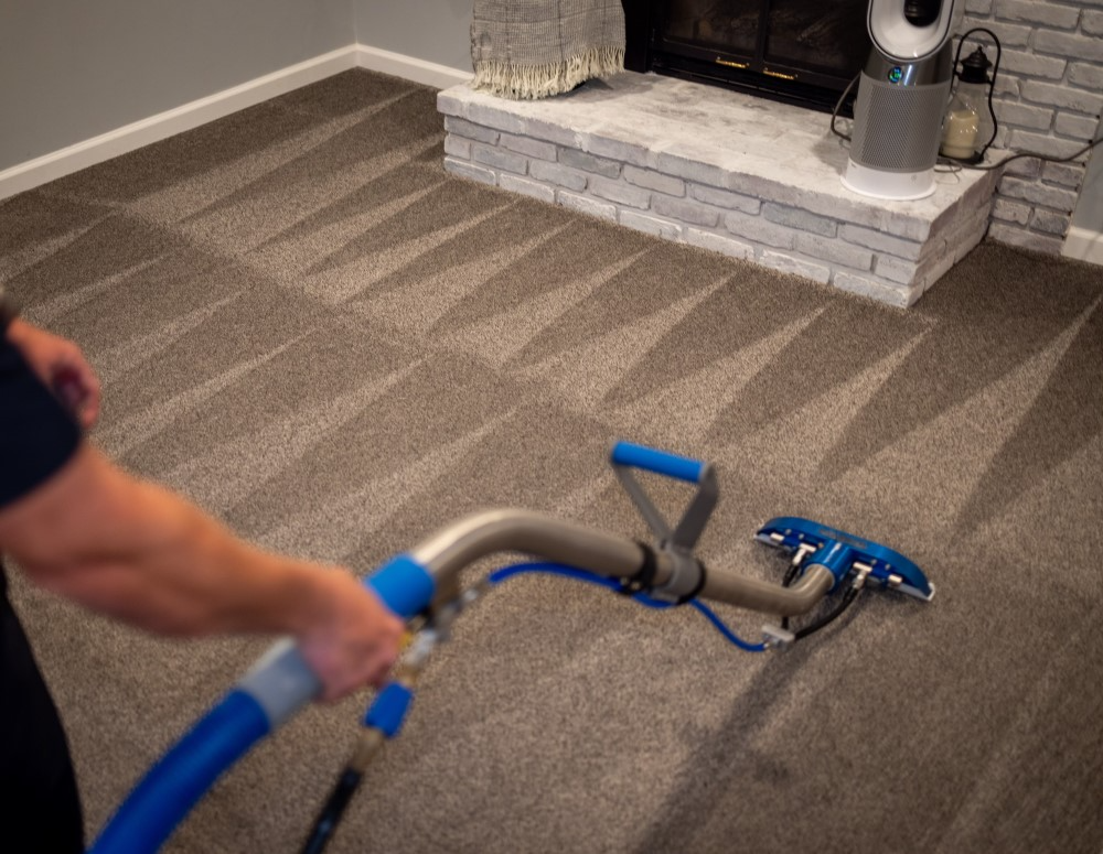 Person cleaning a patterned carpet with a blue and silver carpet cleaner near a fireplace.