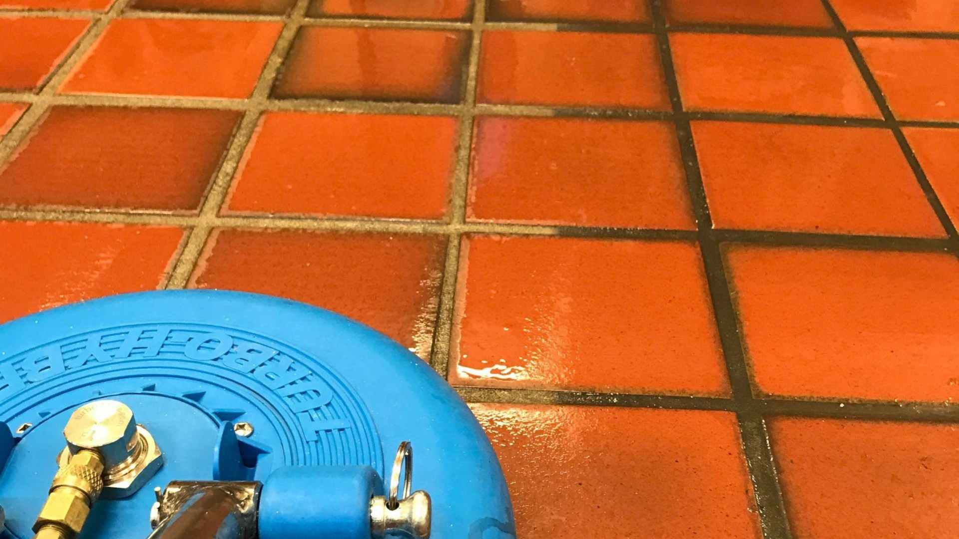 Person using a floor buffer to polish a wet, tiled floor; yellow mop bucket in the background.