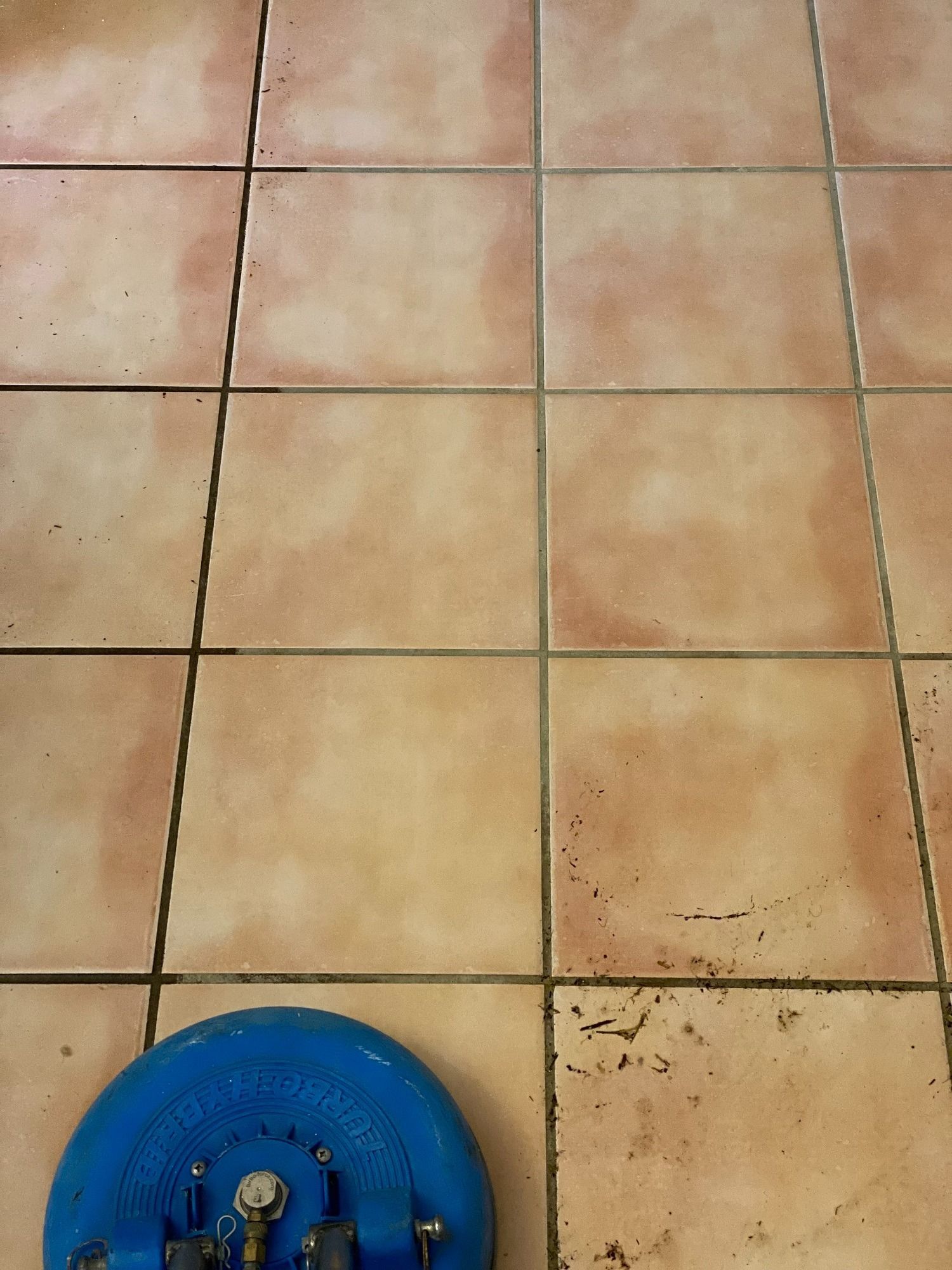 Blue floor cleaning machine scrubbing tan tile floor with dark grout.