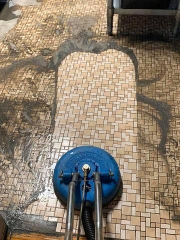 A floor cleaning machine scrubbing a dirty mosaic tiled floor. Dark dirt is being removed.