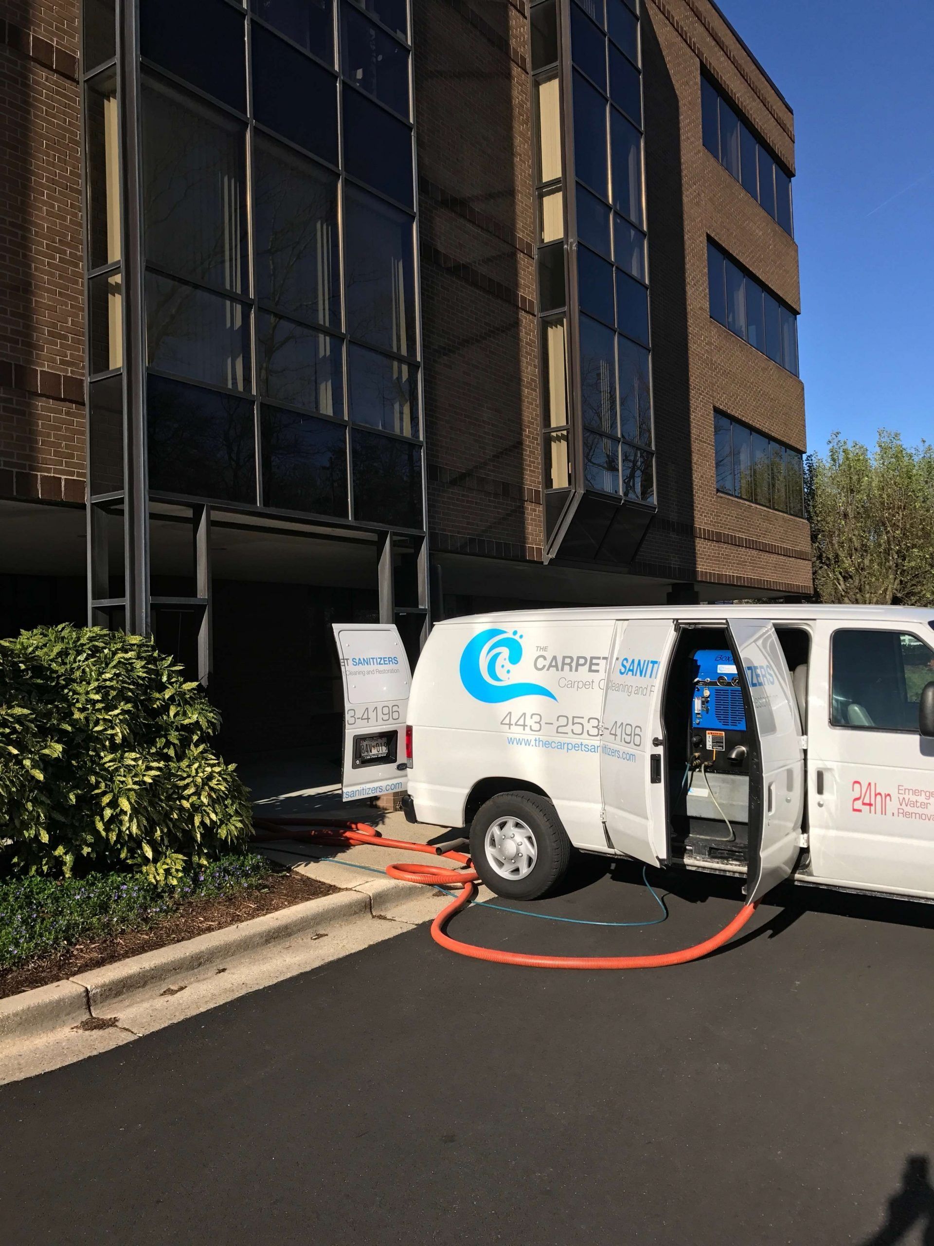 A white van with open doors parked outside a building, orange hose connected for carpet cleaning.