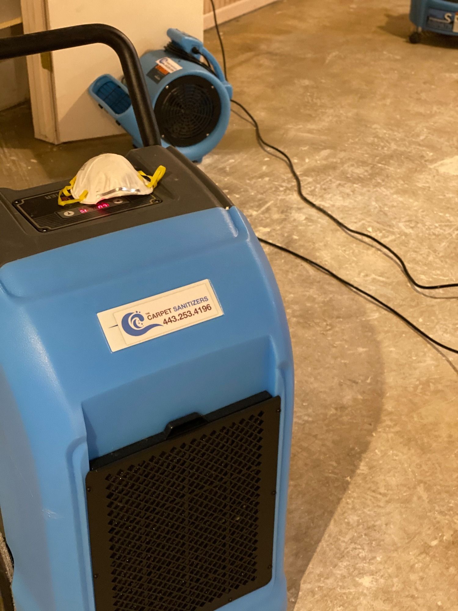 Blue dehumidifier and fan in a room, a face mask rests on the dehumidifier.