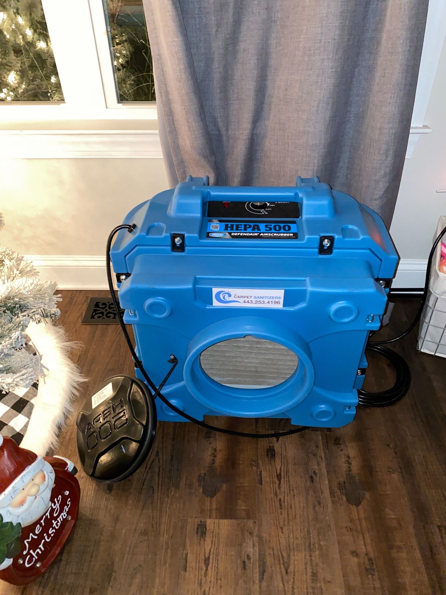 Blue portable air cleaner on a wood floor near a window, curtain, and Christmas decorations.