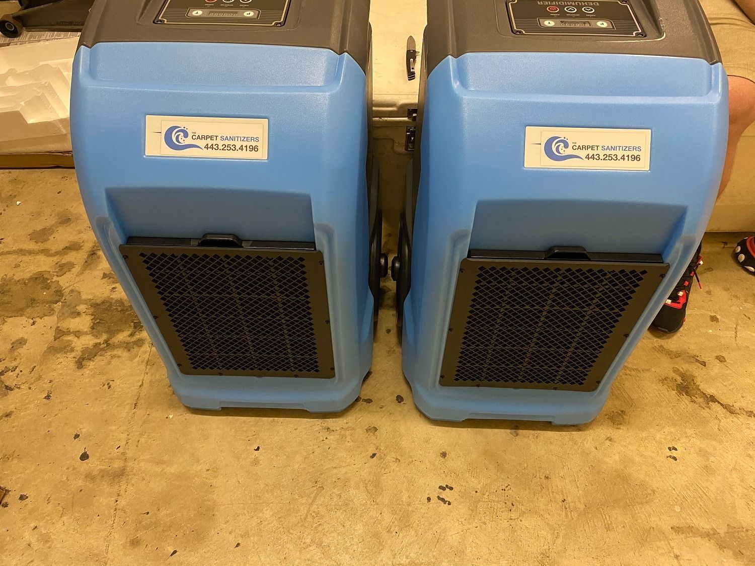 Two blue and gray dehumidifiers side-by-side on a concrete floor with black vents.