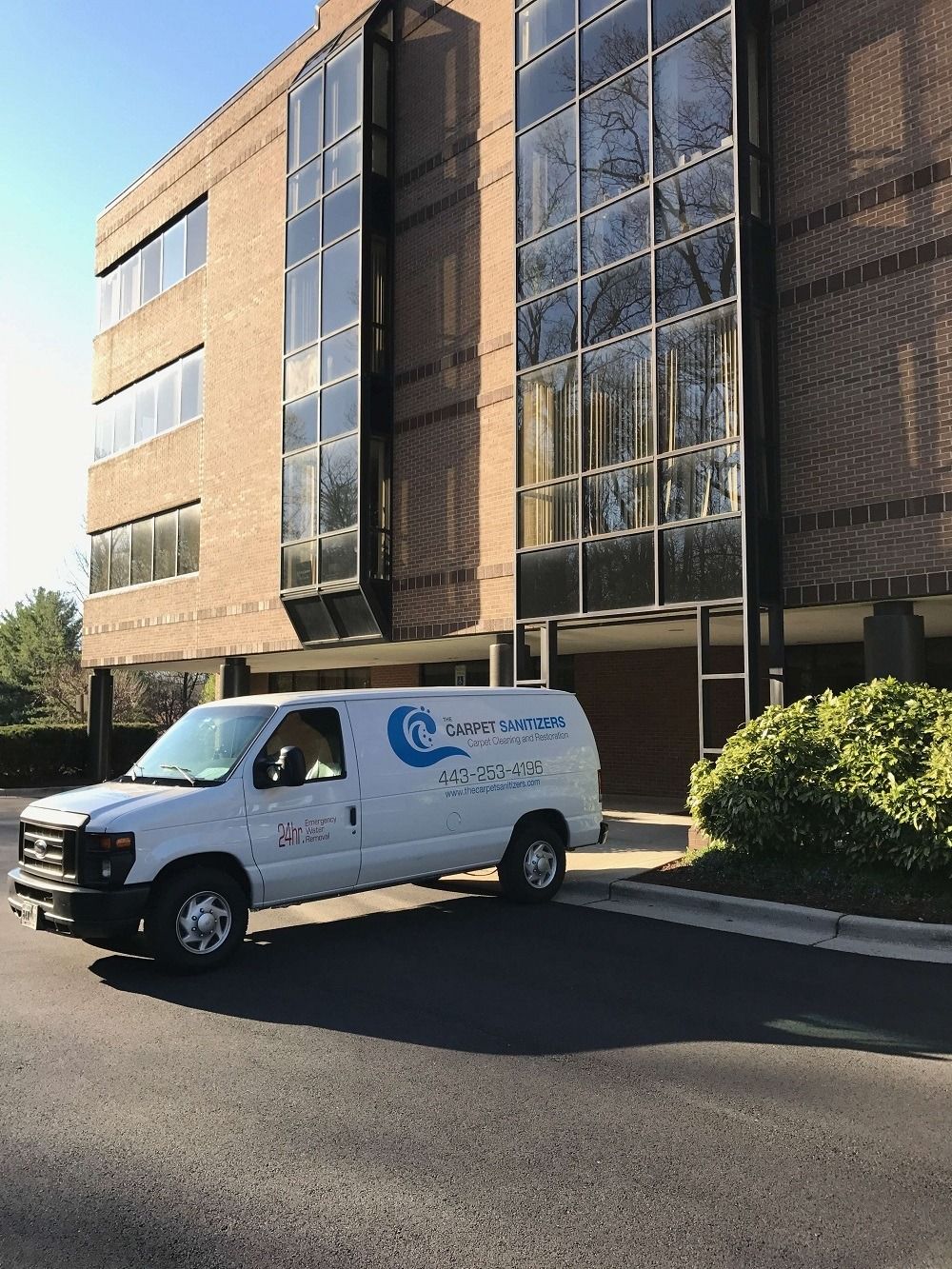 White van with logo parked in front of brick building with glass windows.
