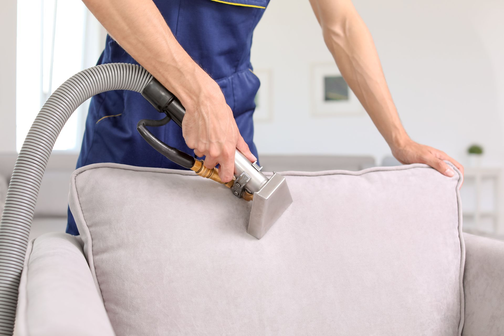 Person vacuuming a light gray upholstered couch with a handheld cleaning tool.