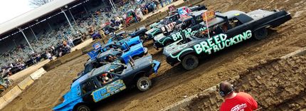Damaged car at Spring Explosion, green and black metal, muddy tires, event banner, 