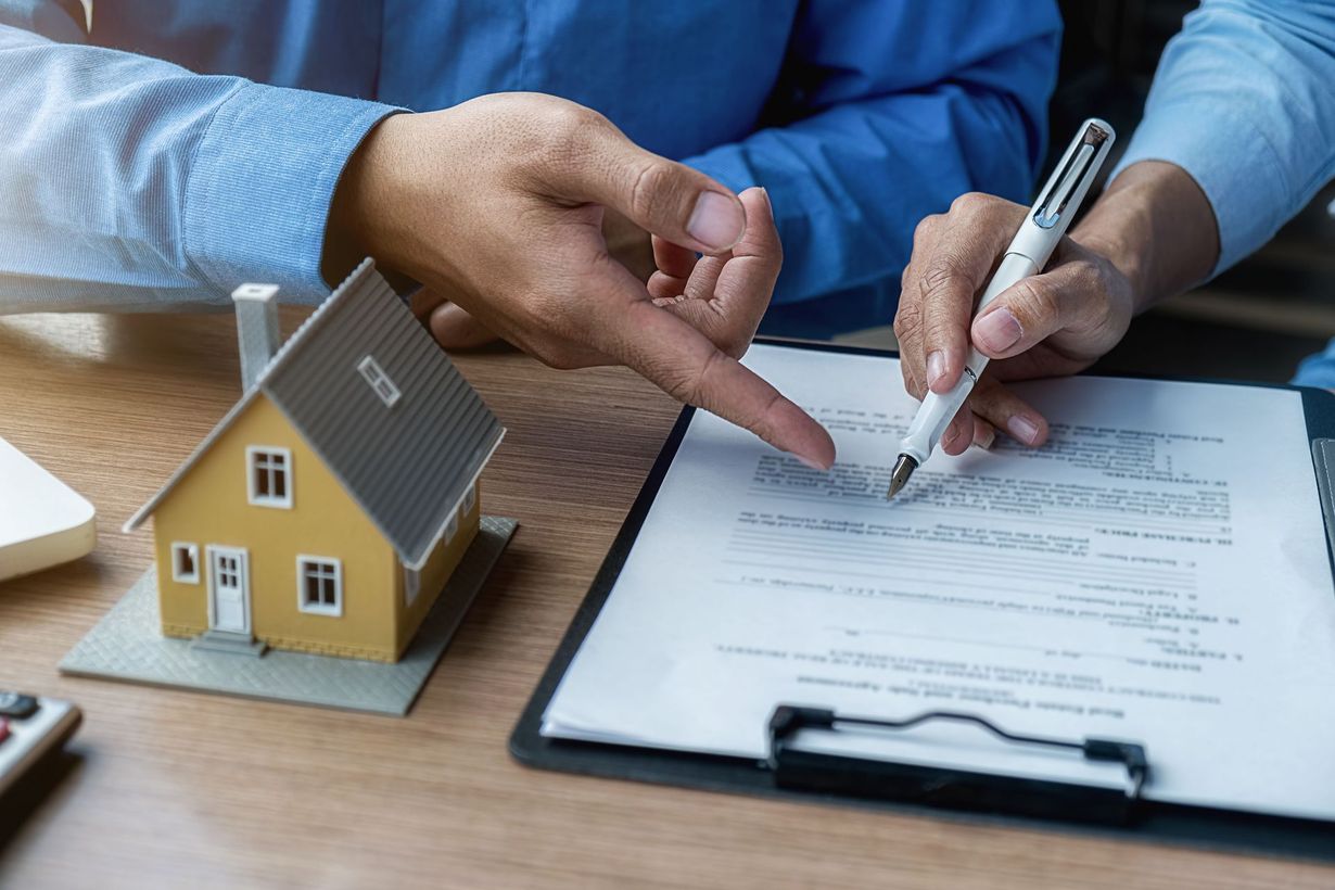 Two people are reviewing a document at a desk with a small model house. One is pointing to the paperwork.