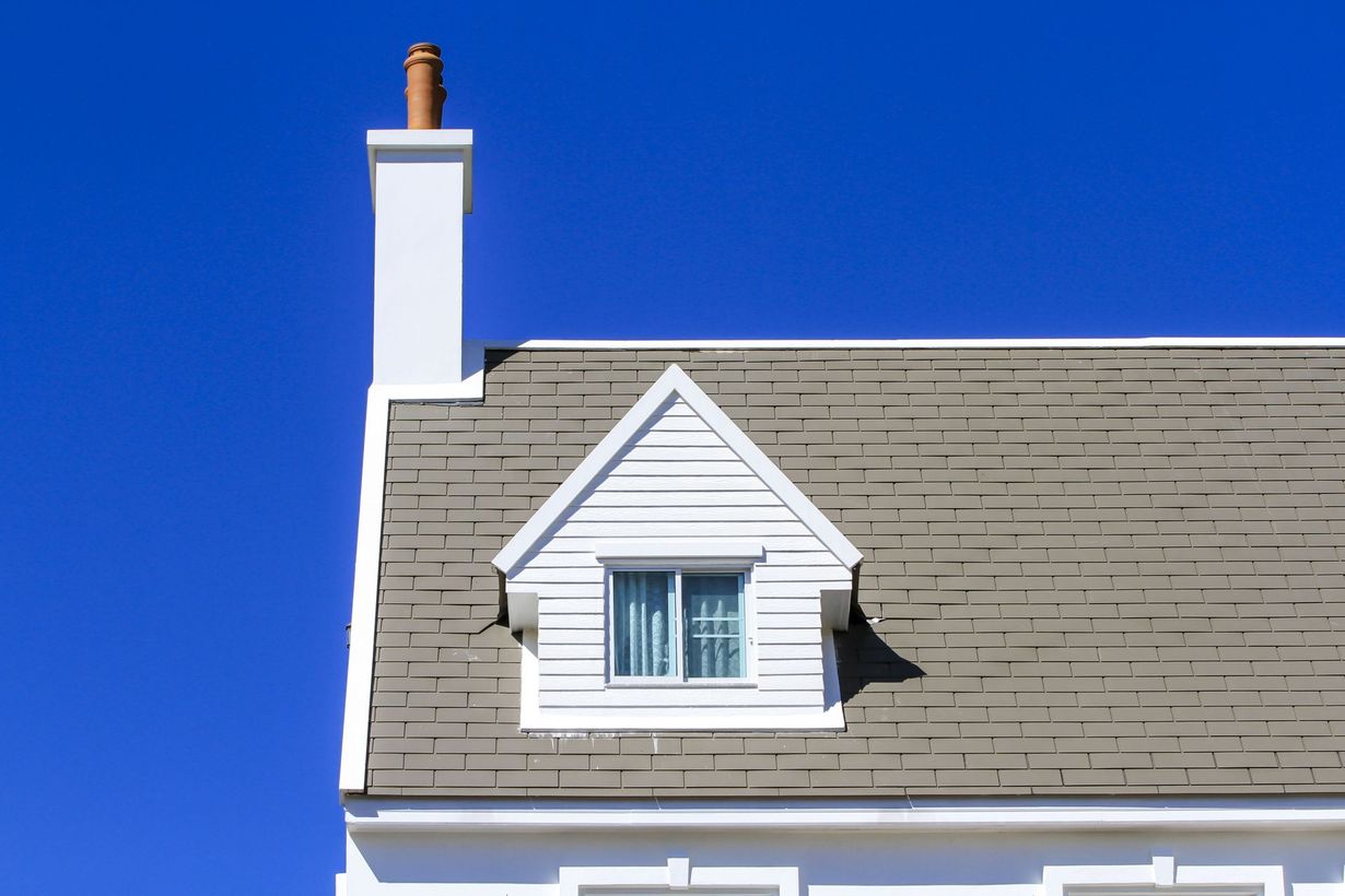 A white chimney and a white-framed dormer window with a grey shingled roof against a solid blue sky.