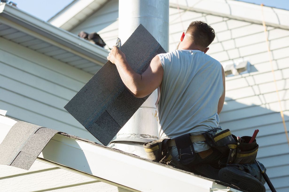 A worker in a grey tank top and tool belt holds a roofing shingle near a metal chimney pipe on a house roof.