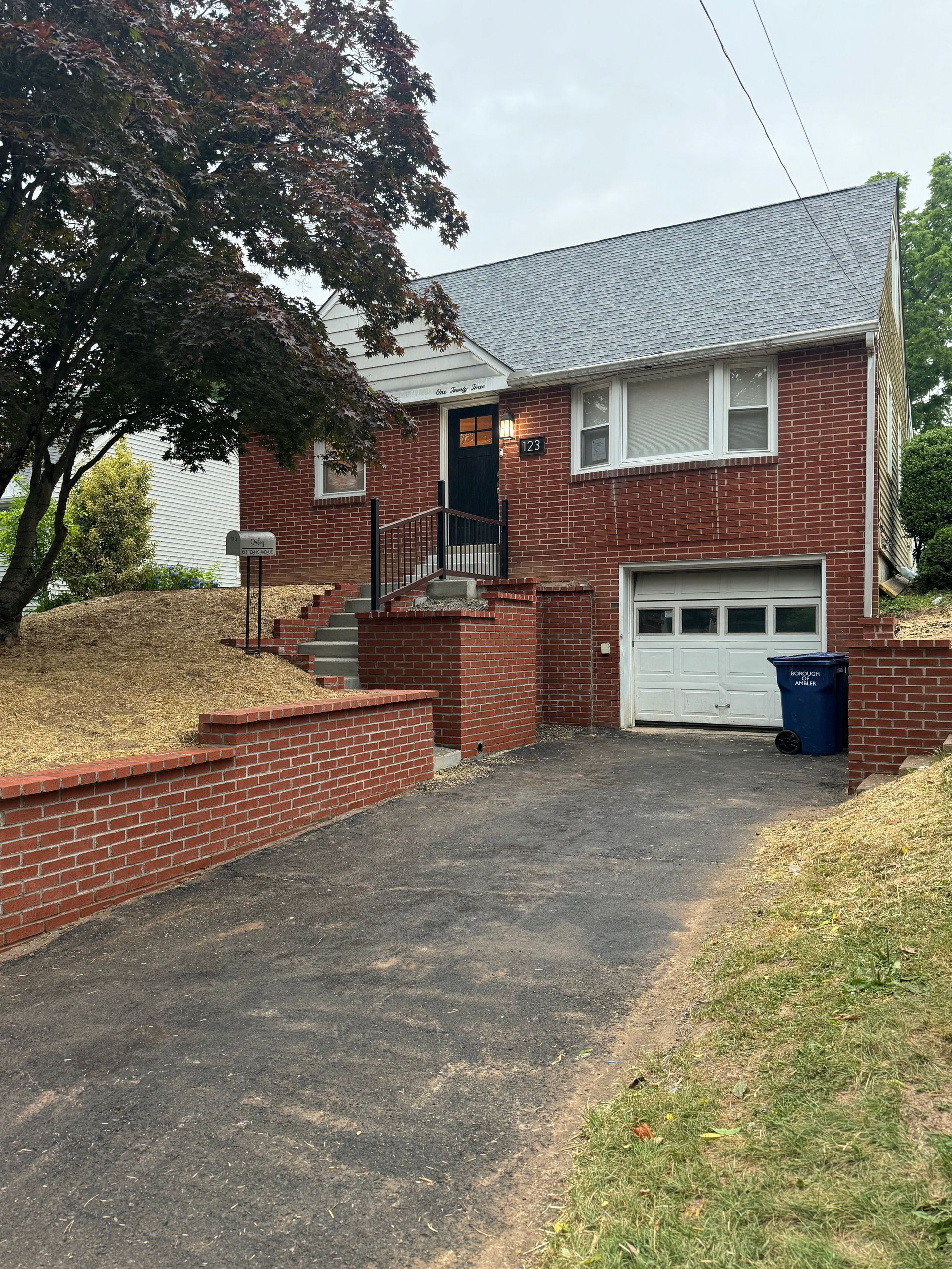 A brick house with a white garage door and a blue trash can in front of it.