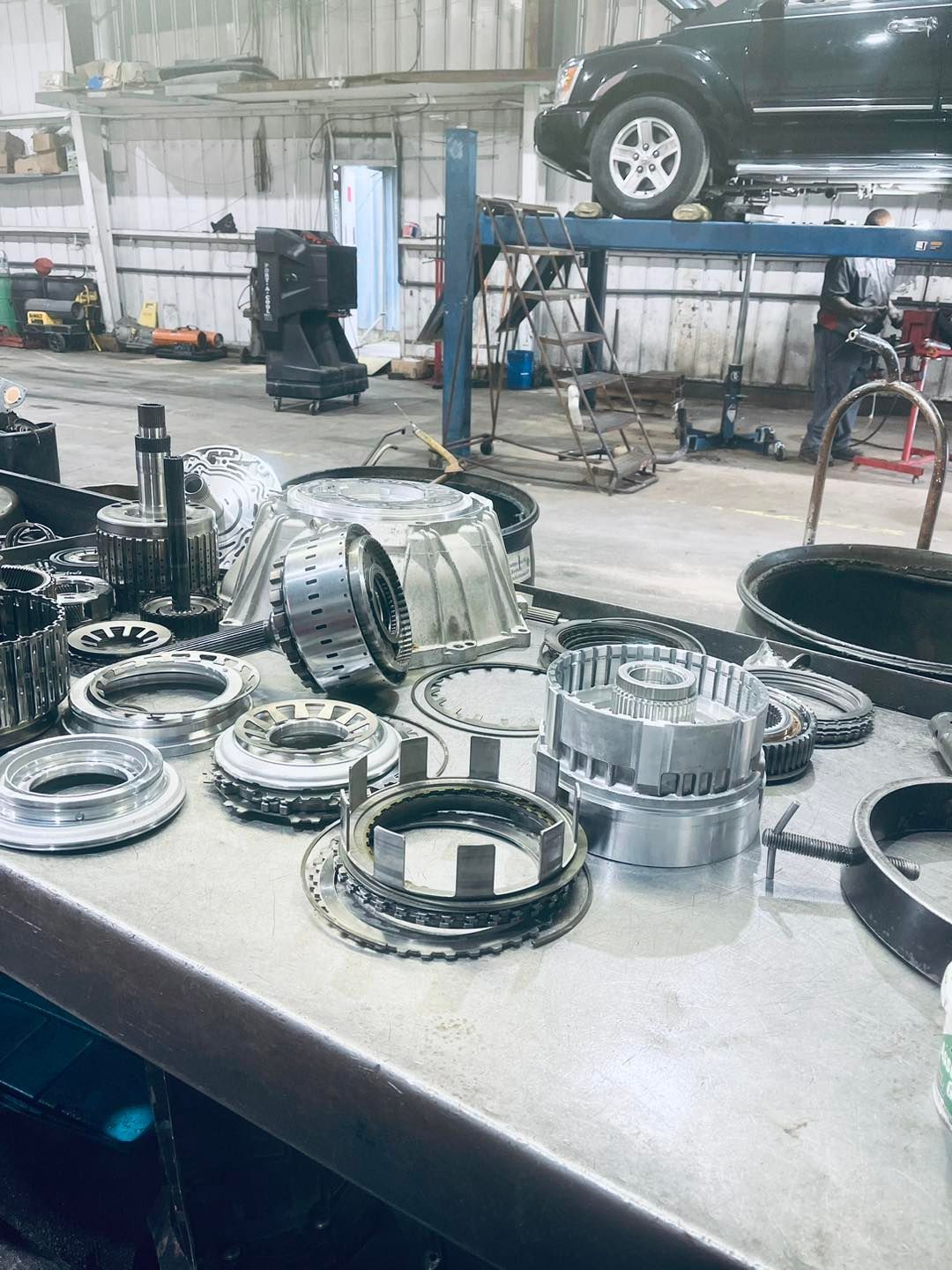 A table with a bunch of gears on it in a garage with a car in the background.