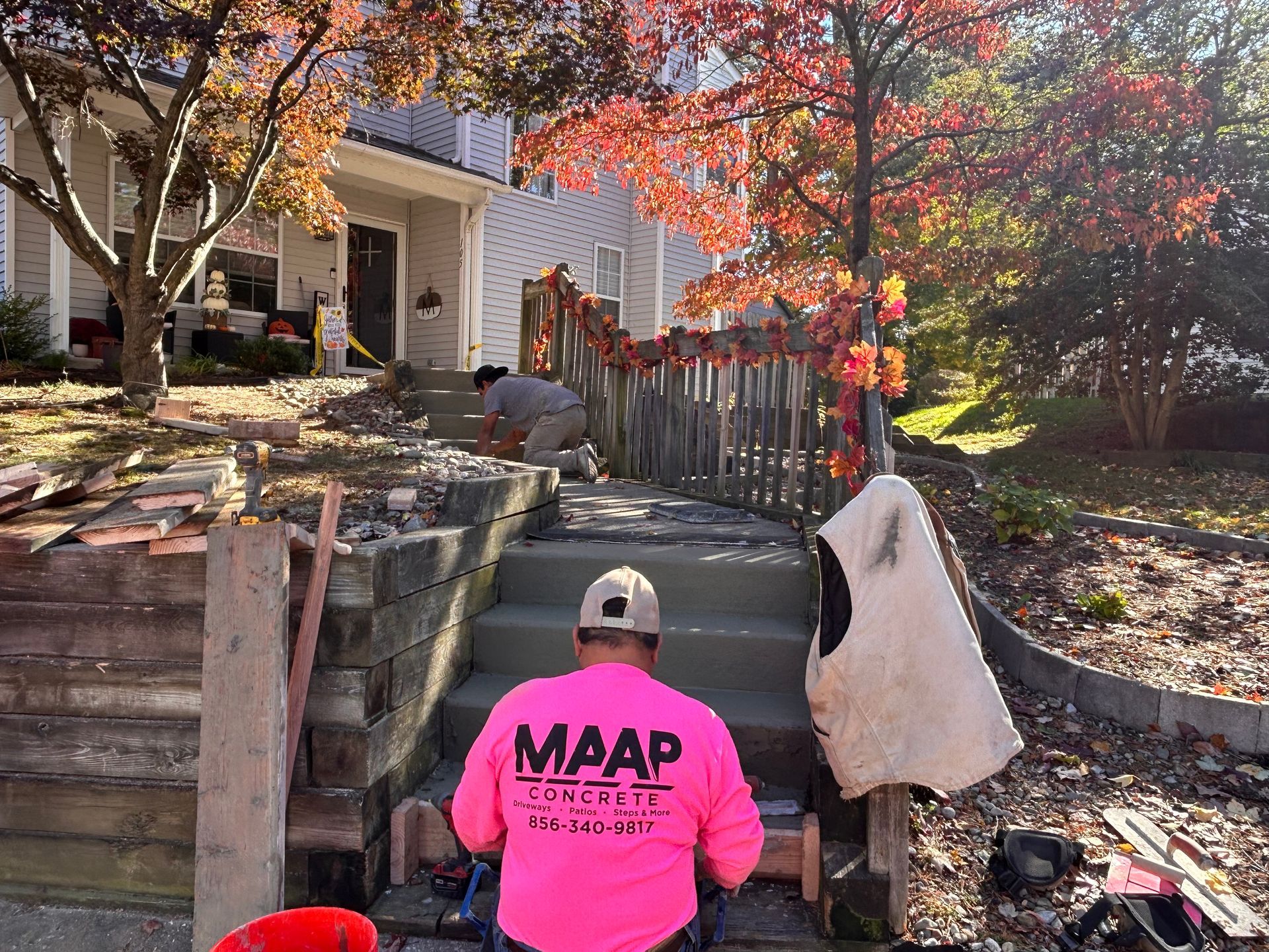 Construction workers resurfacing concrete steps in front of a house. One worker in pink shirt, fall foliage.