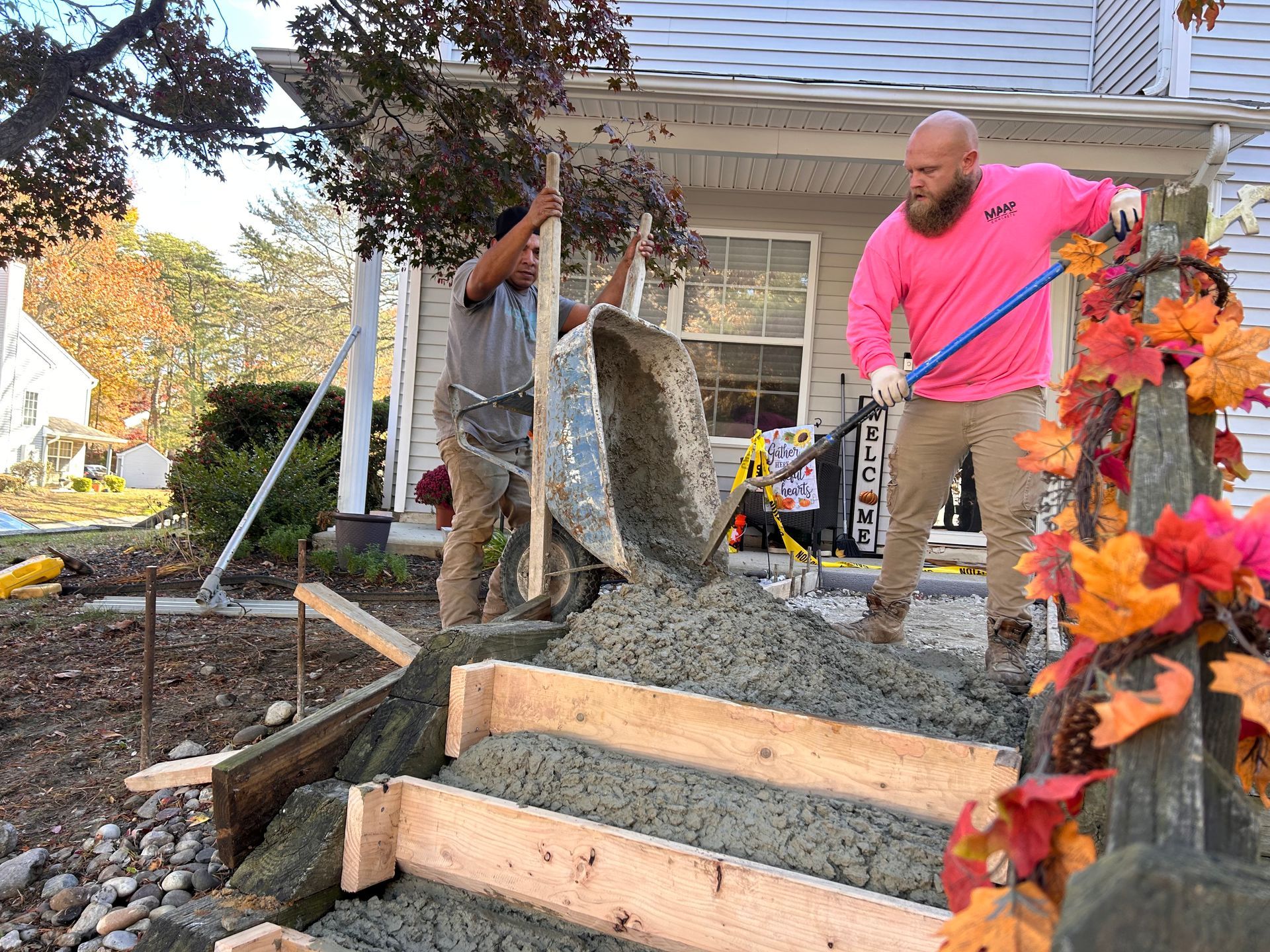 Two workers pouring concrete into wooden forms for stairs on a house's porch.