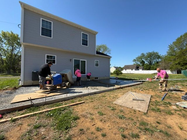 People working outside a gray house, installing or repairing a walkway in a sunny yard