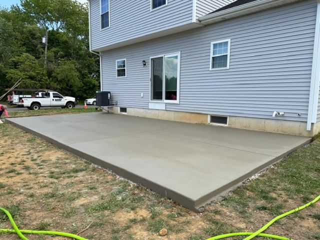 Fresh concrete patio beside a gray house, with a driveway and truck in the background.